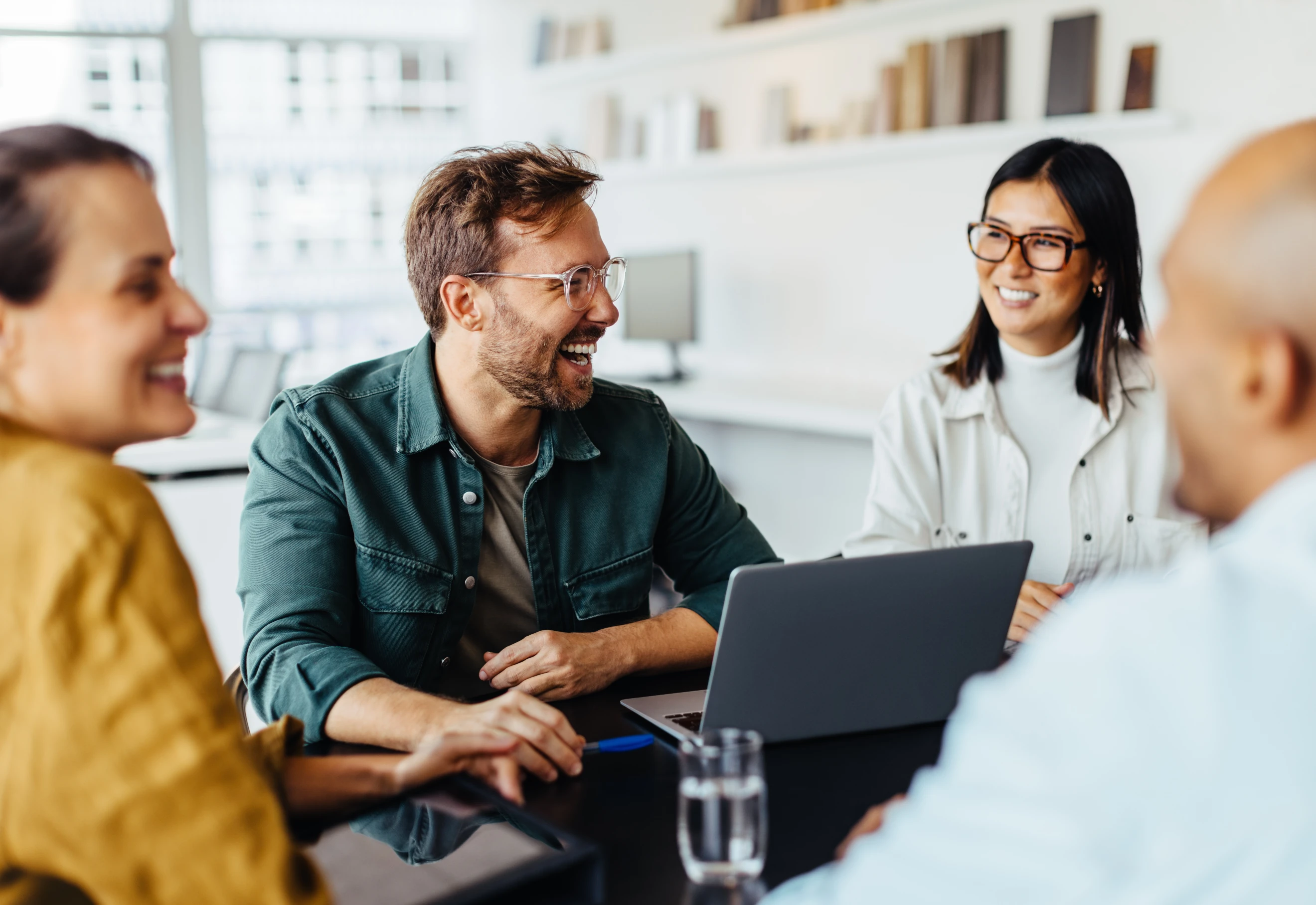 Team of two men and two women laughing at a table with a laptop on it in discussion.